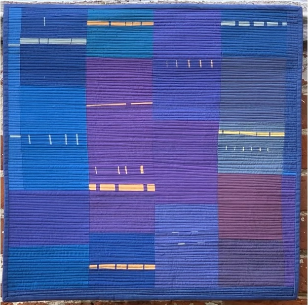 A square quilt with a predominantly blue background. The quilt features a patchwork design with rectangles in various shades of blue and purple. Some rectangles have thin, horizontal lines stitched onto them, and there are small, yellow rectangular shapes scattered throughout. The background is quilted with horizontal lines.