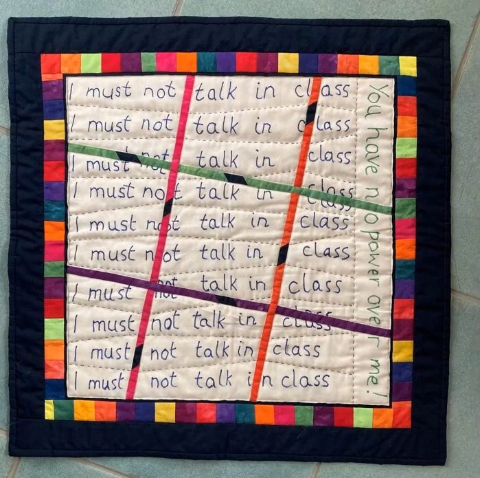 A square quilt with a dark blue background. The quilt features the phrase "I must not talk in class" repeated multiple times in a grid-like pattern. The phrase is written in colorful letters, with each letter made up of small, colorful squares. The background is quilted with a linear pattern. The quilt has a colourful border with the phrase "You have no power over me!" written in white letters.