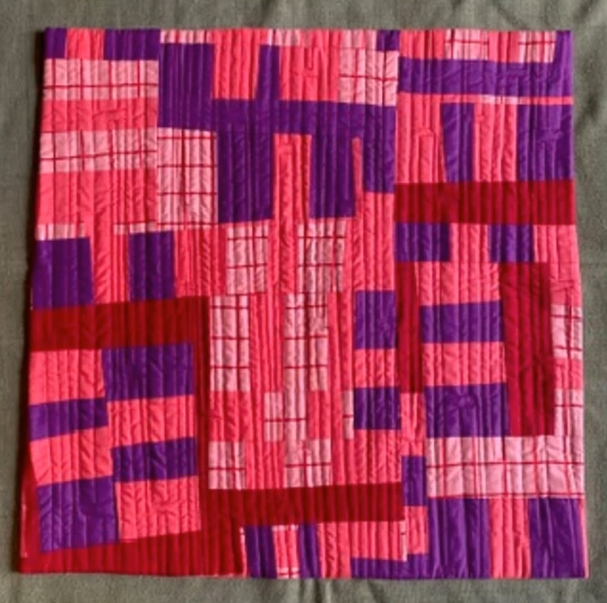 A rectangular quilt with a vibrant, geometric design. The quilt features a patchwork of pink and purple squares and rectangles, with some areas textured with vertical lines. The colours create a striking contrast and the overall design is visually dynamic.