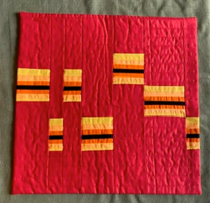 A rectangular quilt with a vibrant pink background. The quilt features multiple rectangular shapes in shades of orange and yellow, with black stripes. The background has a textured, wavy pattern.