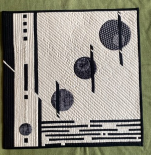 A square quilt with a white background. The quilt features three black circles with a textured pattern, arranged diagonally. Black lines intersect the circles and form a grid-like pattern on the background. The quilt has a black border.