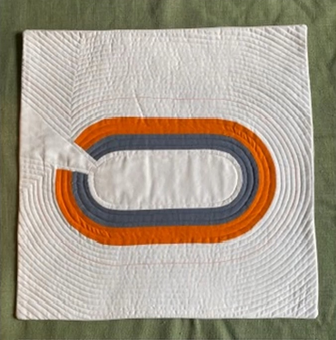 A square quilt with a white background. The quilt features a series of concentric ovals in orange and gray, creating a sense of depth and dimension. The background is quilted with radiating lines.