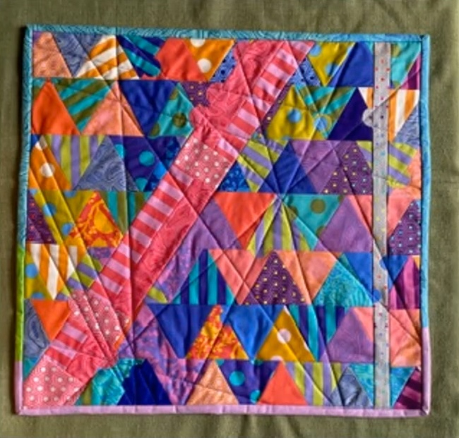 A square quilt with a colorful, geometric design. The quilt is made up of triangles in various colors, including pink, purple, blue, yellow, and orange. The triangles are arranged in a seemingly random pattern, creating a dynamic and visually interesting composition. The quilt has a multicolour border.