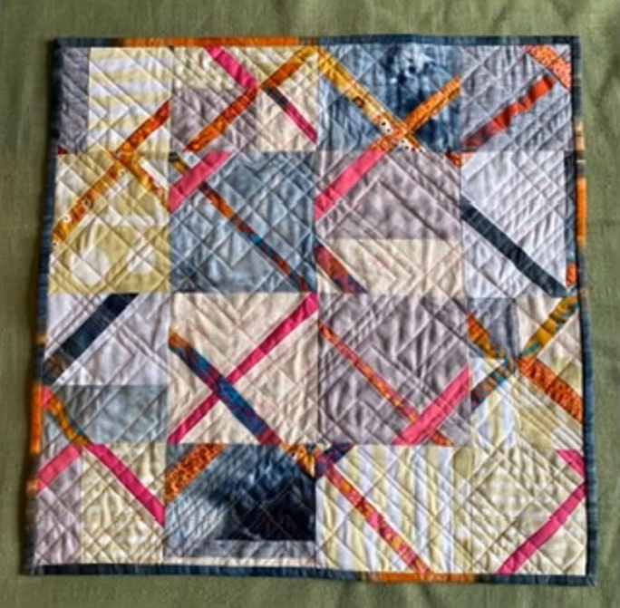 A square quilt with a patchwork design. The quilt features squares in various colors and patterns, including gray, yellow, pink, and orange. There are also diagonal lines of fabric intersecting the squares, creating a dynamic composition.