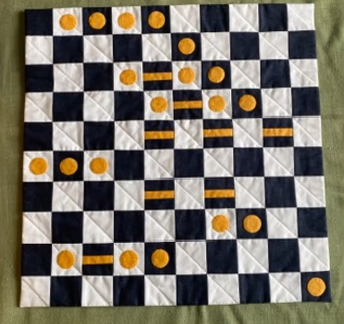 A square quilt with a black and white checkered pattern. There are yellow circles placed on some of the squares, resembling a board game or puzzle. The background is quilted with a linear pattern.