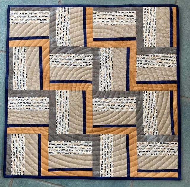 A square quilt with a predominantly beige background. The quilt features a geometric design with intersecting rectangles and curved lines in shades of orange, blue, and gray. The background is quilted with a linear pattern.