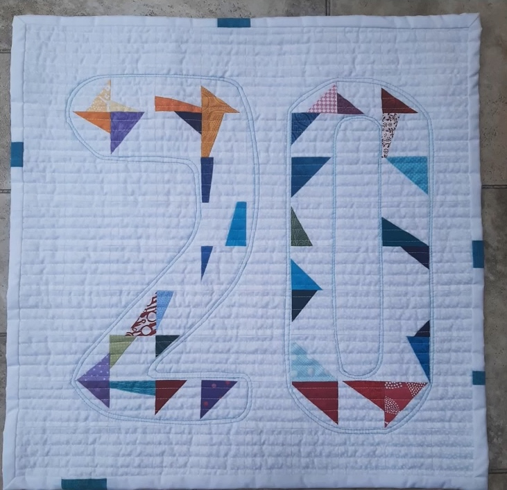 A square quilt with a white background. The numbers "20" are made up of colorful patchwork triangles. The background is quilted with a linear pattern.