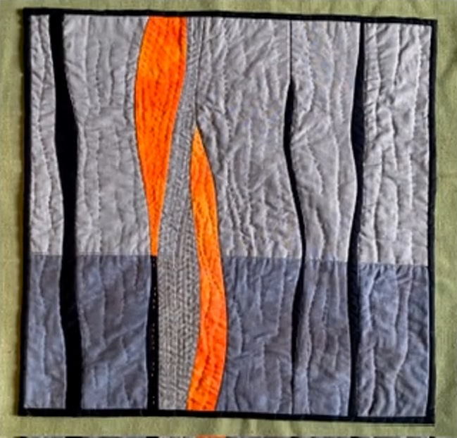 A rectangular quilt with a gray background. The quilt features vertical, wavy lines in shades of gray, with two bright orange lines in the centre. The background is quilted with a subtle, linear pattern. The quilt has a black border.