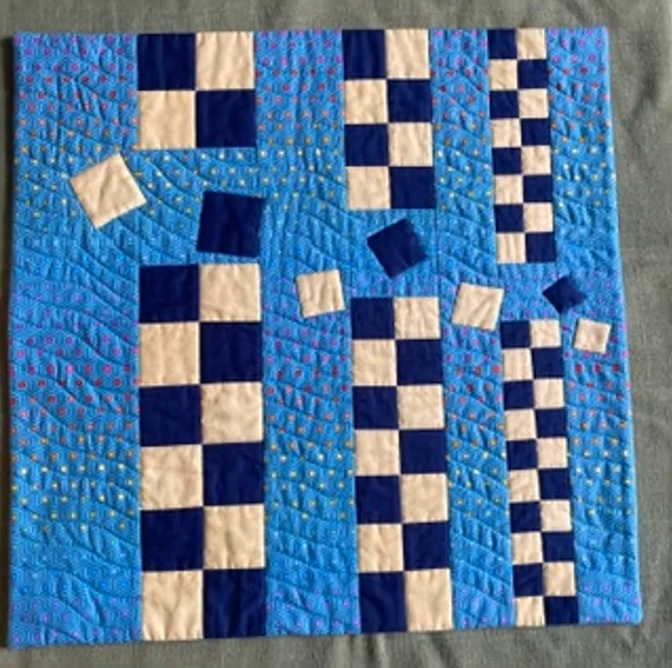 A square quilt with a blue background. The quilt features a patchwork design with squares in blue and white, arranged in vertical columns. There are also smaller, scattered squares in various sizes and patterns. The background is quilted with a curved line.