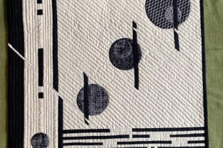 A square quilt with a white background. The quilt features three black circles with a textured pattern, arranged diagonally. Black lines intersect the circles and form a grid-like pattern on the background. The quilt has a black border.