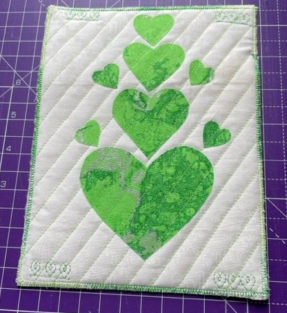 A square quilt with a white background. The quilt features a series of green hearts, arranged in a pyramid shape. The hearts are made of fabric with a marbled pattern. The background is quilted with a diagonal line pattern. The quilt has a green border.