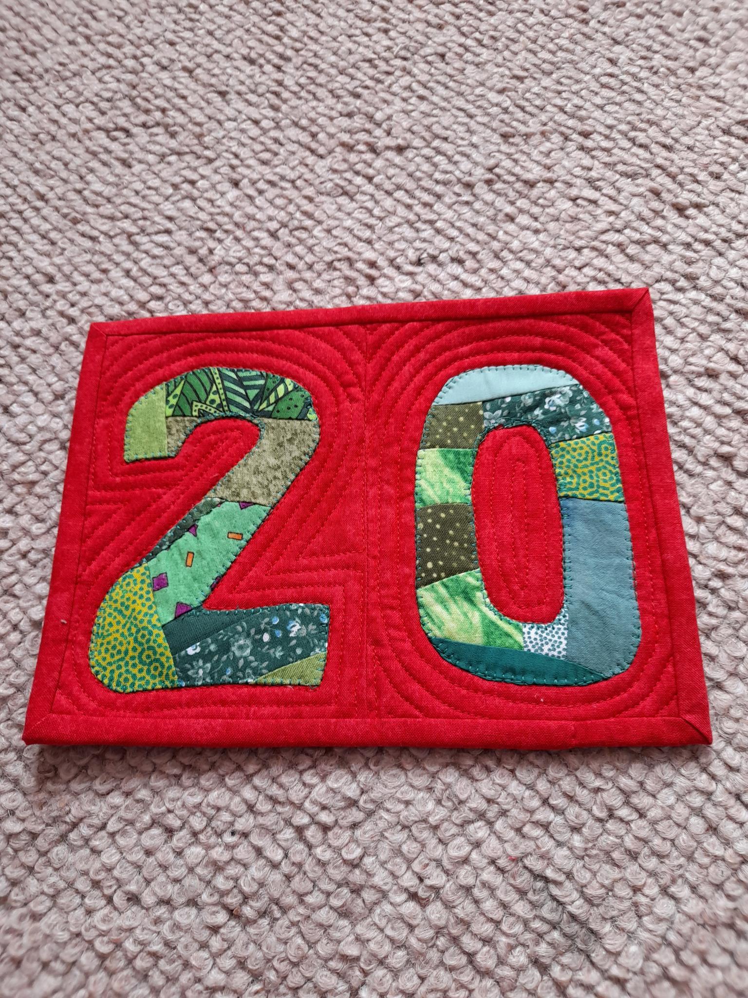 A rectangular quilt with a red background. The numbers "20" are made up of patchwork fabric in shades of green and blue. The background is quilted with echoing the numbers.
