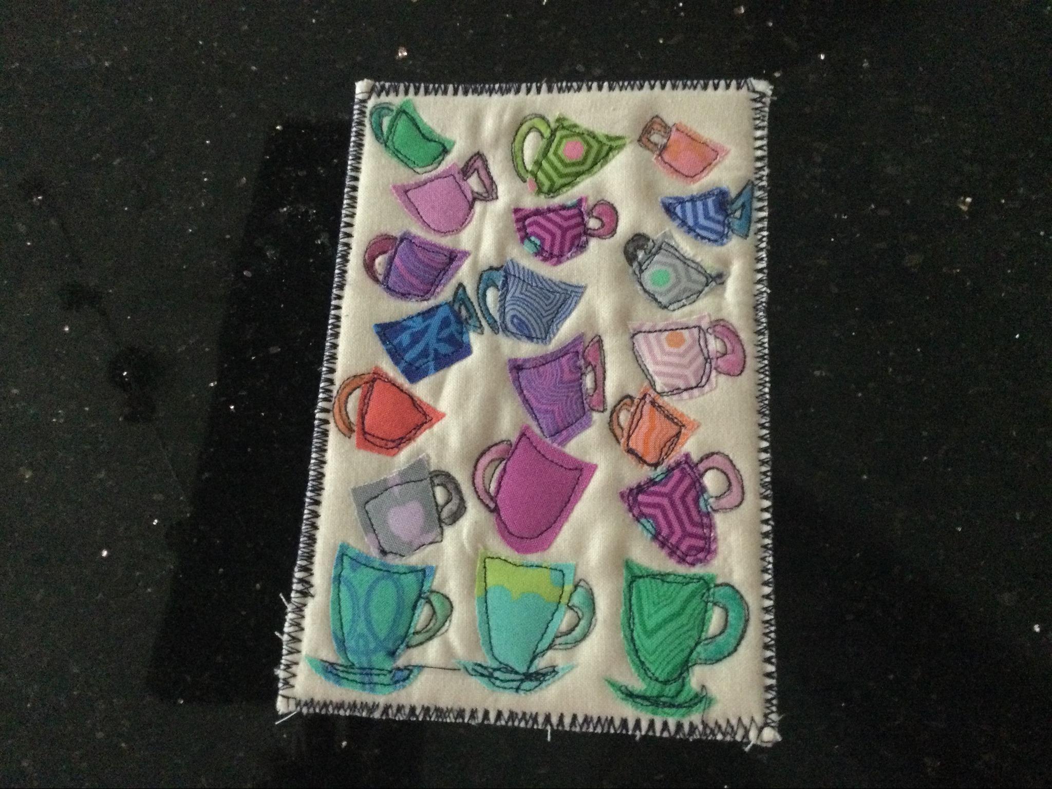A rectangular quilt with a white background. The quilt features multiple colourful mugs in various shapes and patterns, scattered across the background. The mugs are made of different fabrics and have unique designs. The background is quilted with a subtle, linear pattern. The quilt has a black zigzagged stitched border.