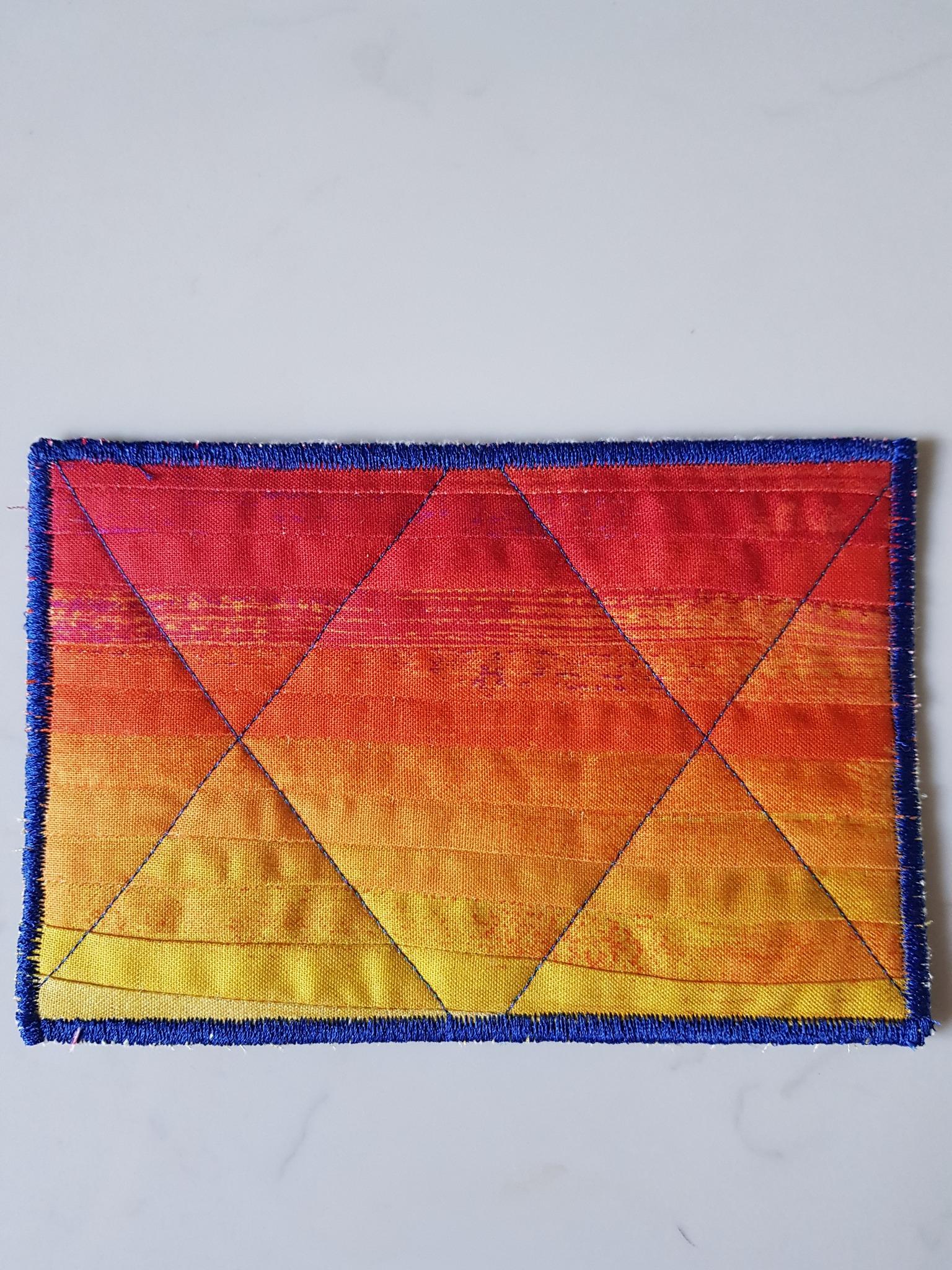A rectangular quilt with a vibrant gradient background, transitioning from red to orange and yellow. The quilt is quilted with a diamond pattern and has a blue border.