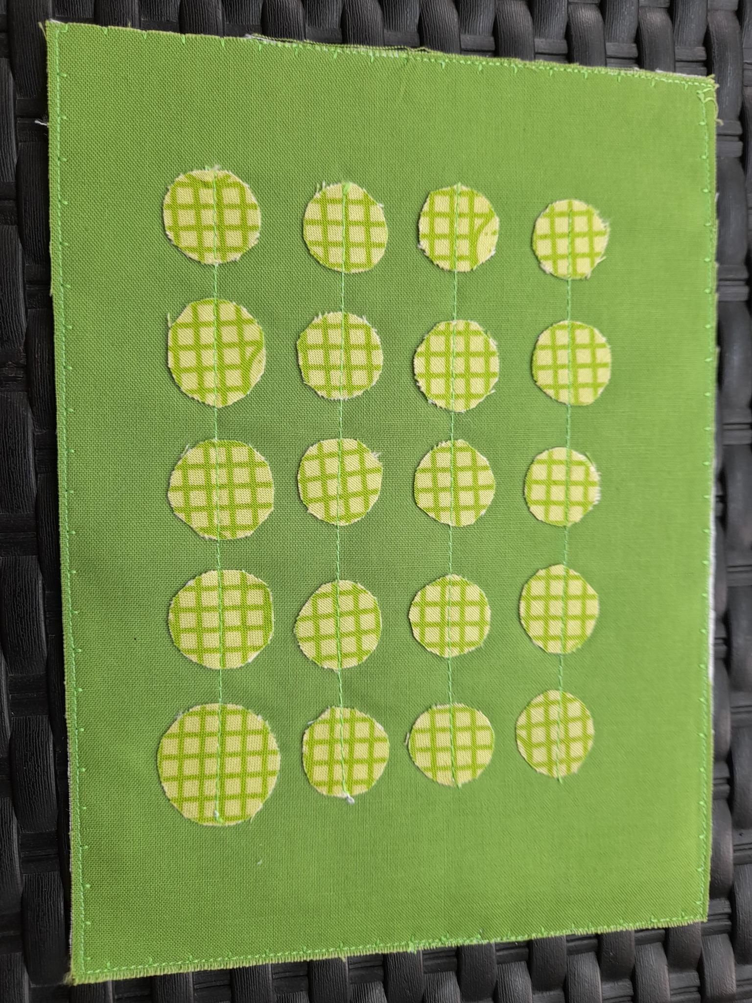 A square quilt with a green background. The quilt features a pattern of yellow oval shapes, each with a grid-like pattern. The shapes are evenly spaced and create a visually pleasing, repetitive design. The background is quilted with a subtle, linear pattern.