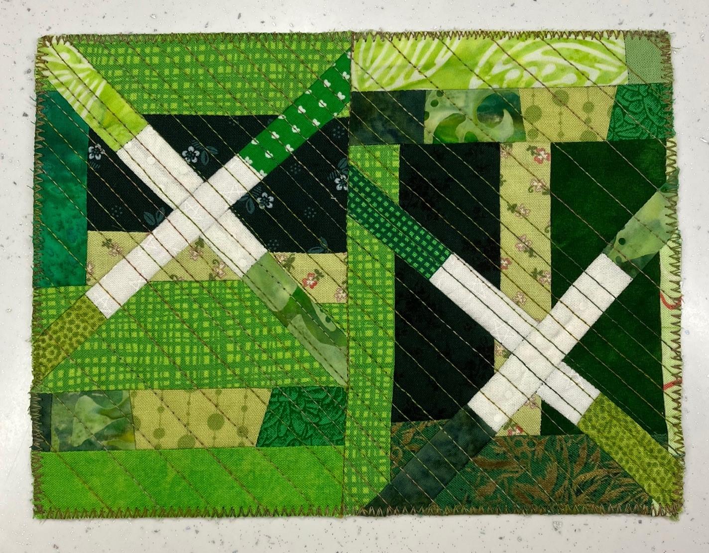 A postcard quilt with a green pieced background. The quilt features a patchwork design with squares, triangles, and rectangles in various shades of green. There are also white lines and shapes, creating a geometric pattern. The background is quilted with a diagonal linear pattern.