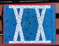 A square quilt with a blue background and a red border. The number "XX" is formed using white strips of fabric, with small, colourful beads scattered around.