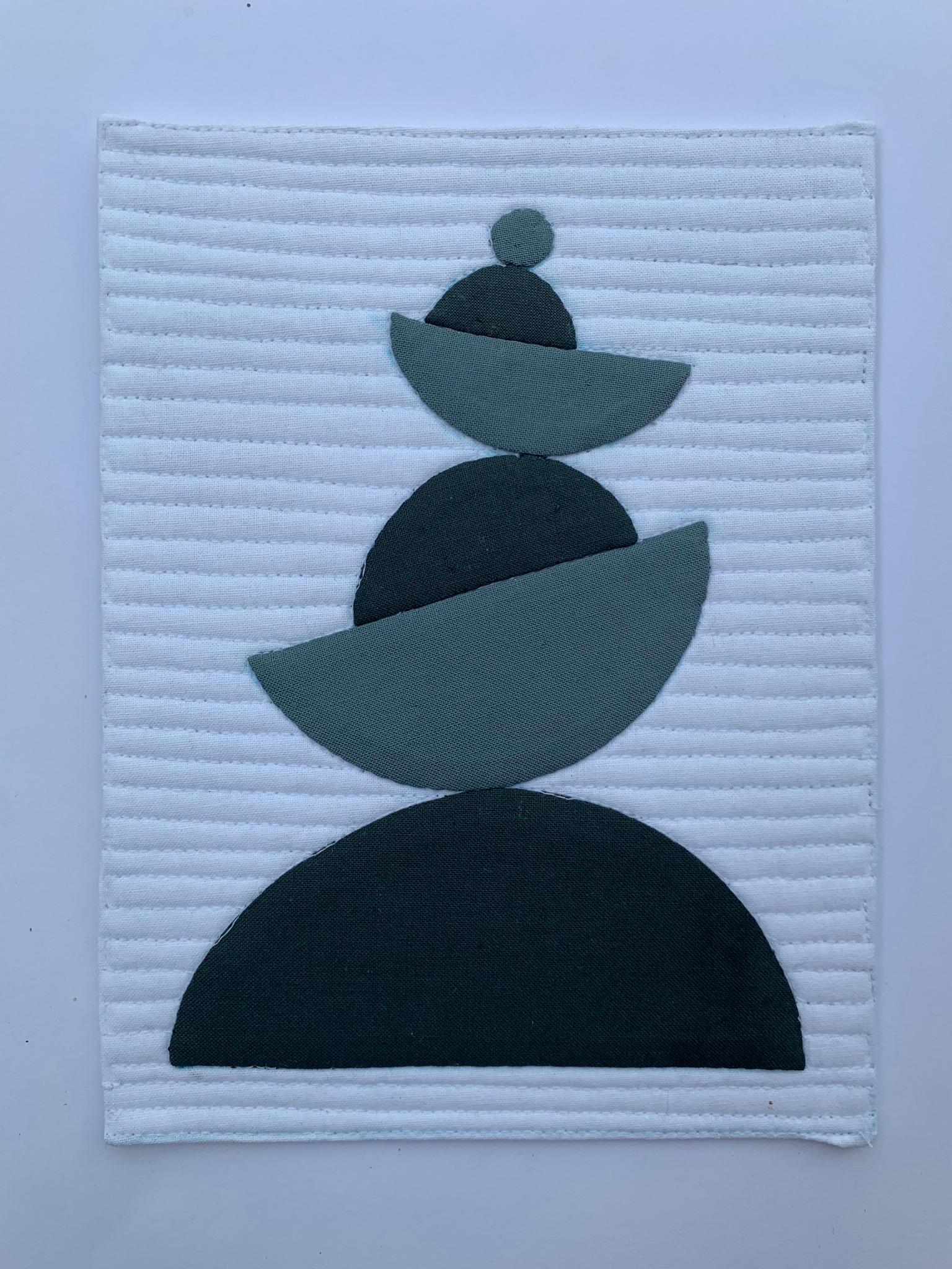 A square quilt with a white background. The quilt features a stack of overlapping semicircles in shades of grey, resembling a balanced sculpture. The background is quilted with horizontal lines.