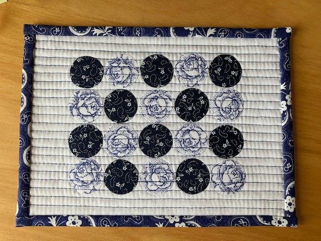 A rectangular quilt with a white background. The quilt features a pattern of overlapping circles in shades of blue, with a floral design on the fabric. The circles are arranged in rows. The quilt has a blue border.
