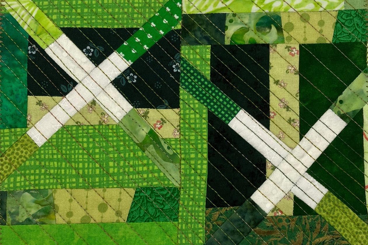 A postcard quilt with a green pieced background. The quilt features a patchwork design with squares, triangles, and rectangles in various shades of green. There are also white lines and shapes, creating a geometric pattern. The background is quilted with