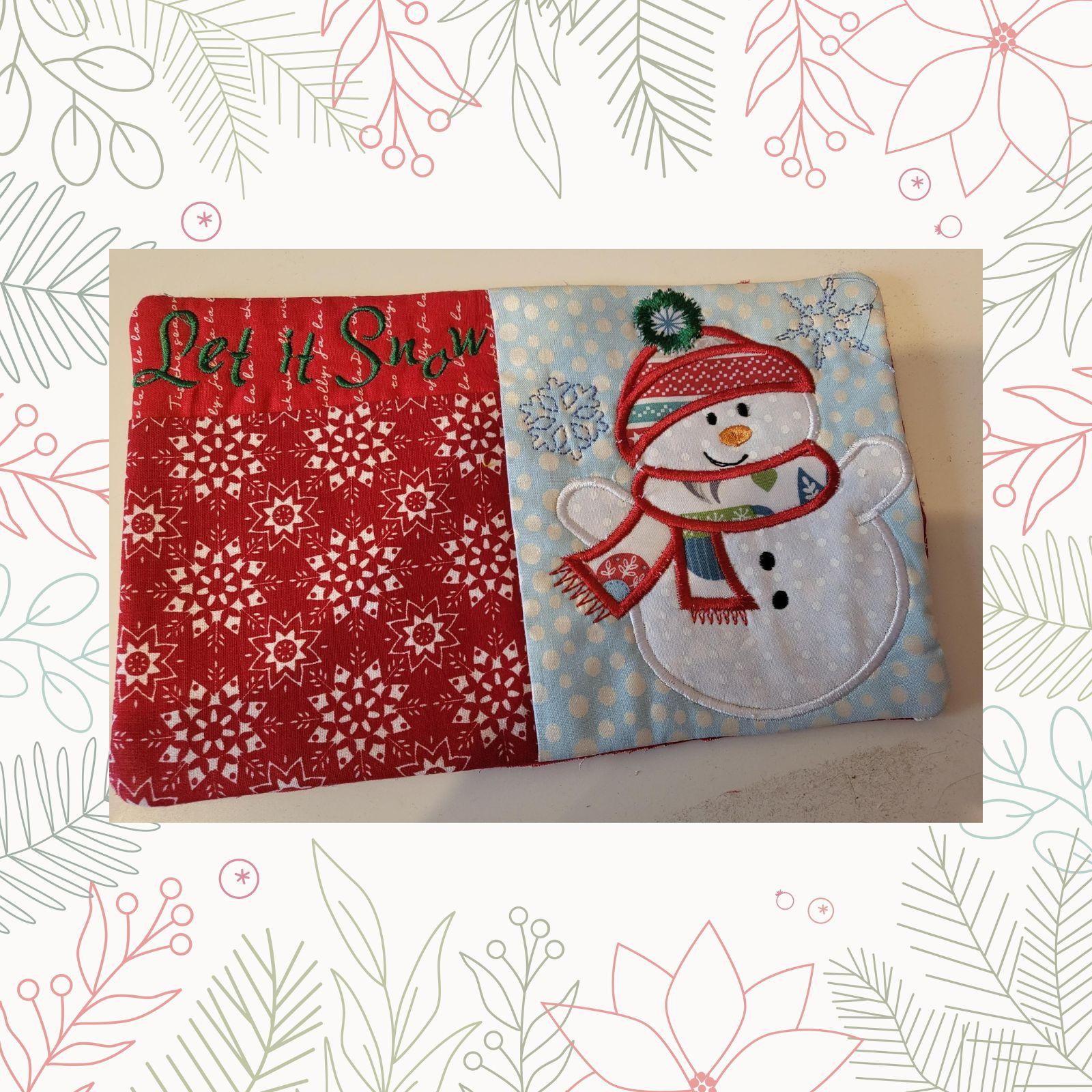 A festive, rectangular quilt with a Christmas theme. The left side features a red background with a snowflake pattern and the phrase "Let It Snow" embroidered in green. The right side depicts a cheerful snowman with a red hat, scarf, and buttons. The snowman is surrounded by snowflakes and a blue sky background.