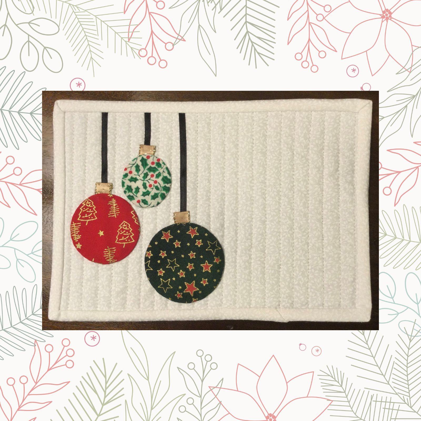 A rectangular quilt with a white background. The quilt features three hanging Christmas ornaments in red, green, and gold. The ornaments are decorated with various patterns, including stars and holly leaves. The background is quilted with vertical lines.