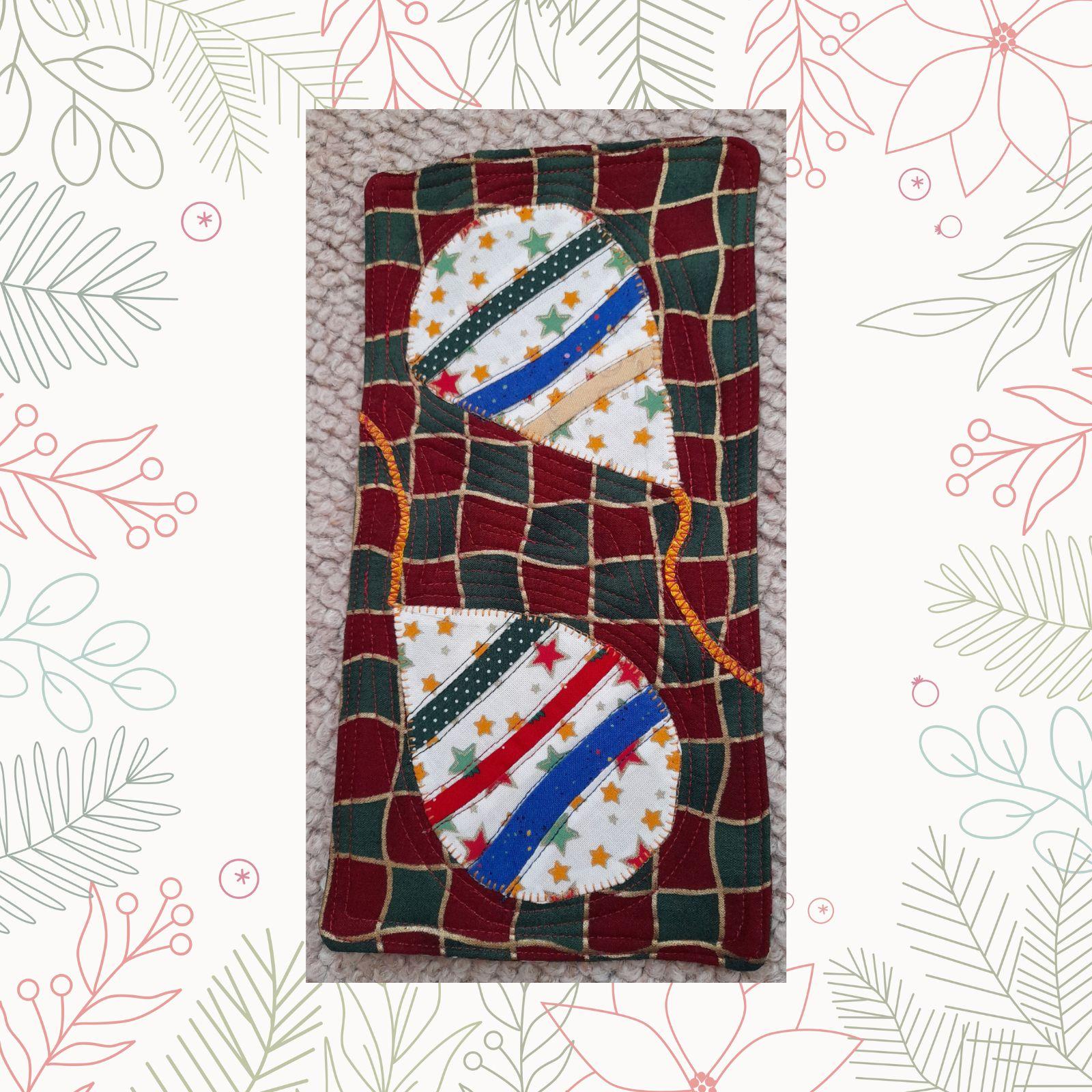 A rectangular quilt with a red background and a green border. The quilt features two teardrop-shaped ornaments, each made from colourful fabric with a pattern of stars.