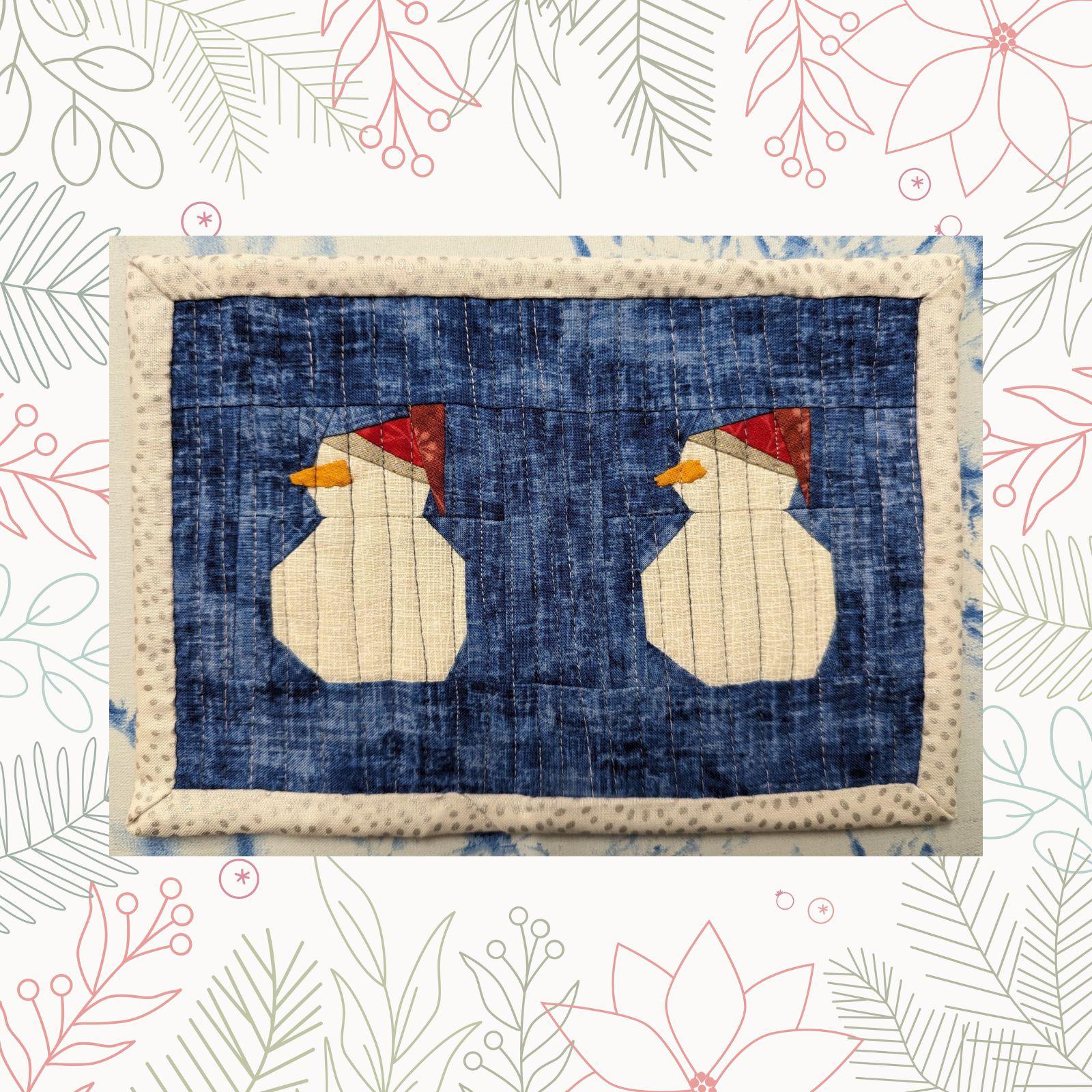 A rectangular quilt with a blue background and a white border. The quilt features two snowmen with red hats, standing side by side. The background is quilted with a linear pattern.