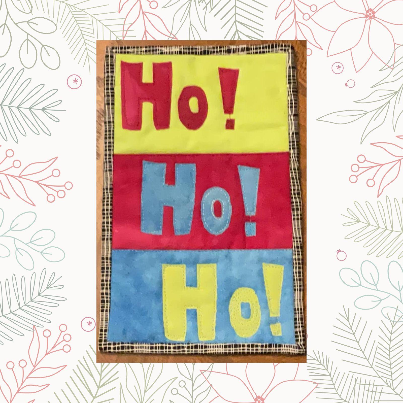 A rectangular quilt with a plaid background and a brown border. The quilt features the word "Ho!" repeated three times in large, colorful letters. The letters are made up of different fabrics in red, yellow, and blue.