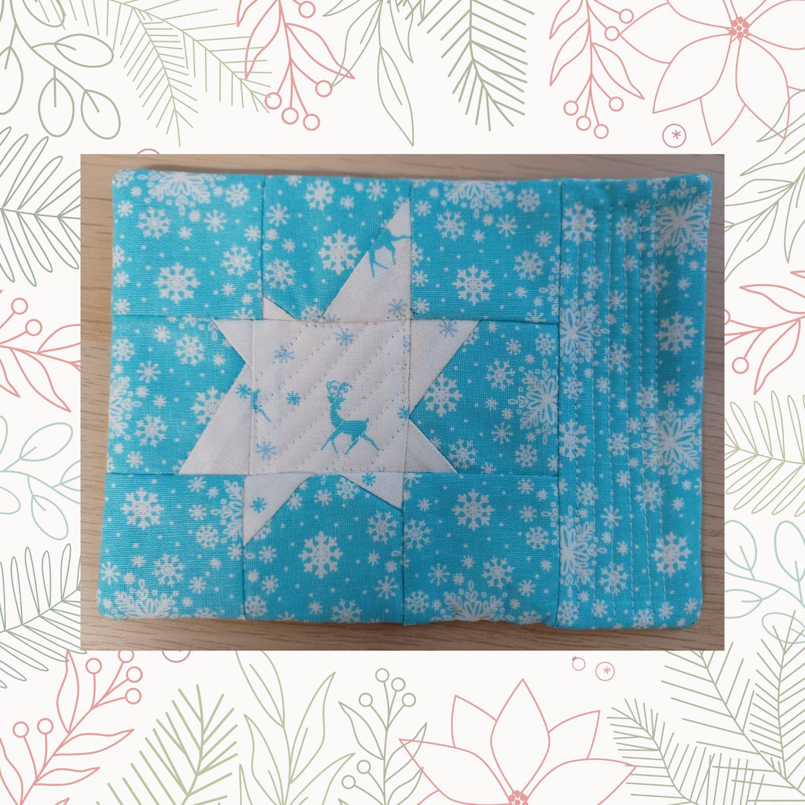 A rectangular quilt with a blue background and a white border. The quilt features a star-shaped design in the center, with a smaller star in the middle. The smaller star has a pattern of snowflakes and a small deer.