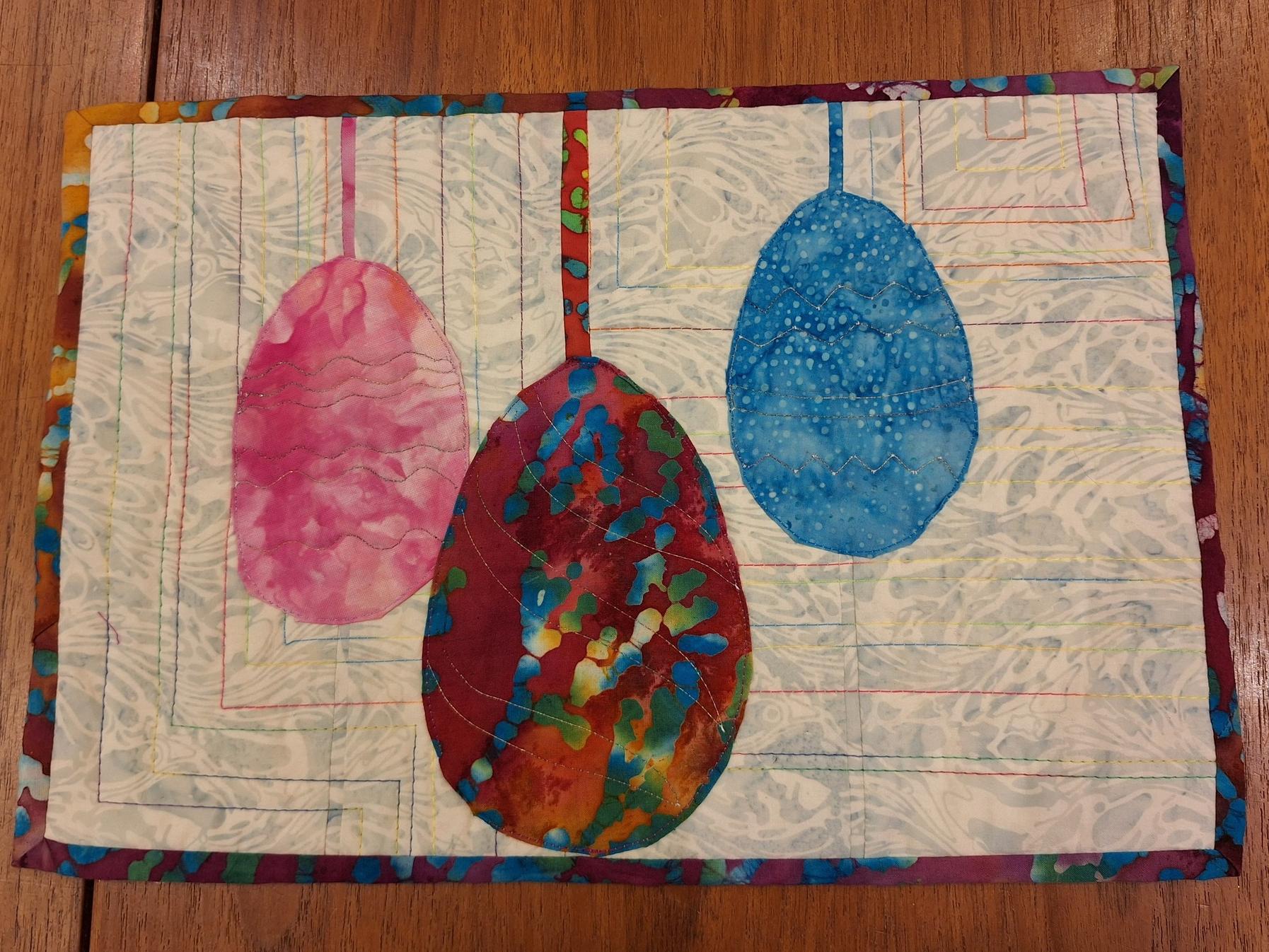 A quilted placemat with a white background and a colourful border. The placemat features three egg-shaped ornaments in pink, red, and blue, hanging from strings. The ornaments are made of batik fabric with intricate patterns. The background is quilted with a linear pattern.