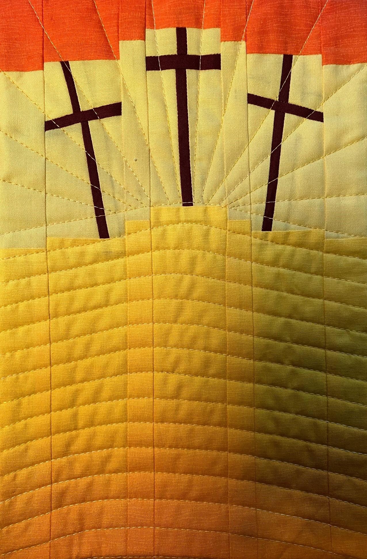 A rectangular quilted placemat with a sunrise-like background. The background transitions from orange to yellow with radiating lines. Three black crosses are positioned on the yellow section, symbolising a religious or spiritual theme. The quilt is quilted with curved lines at the bottom and radiating lines at the top.