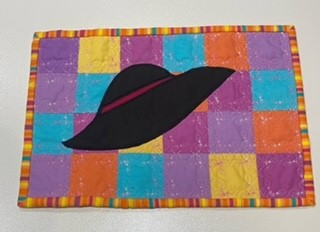 A rectangular quilted placemat with a colourful background and a black hat in the centre. The background is a patchwork of squares in various colours, including purple, yellow, and orange. The hat has a red band around the brim. The placemat is framed by a colourful border.