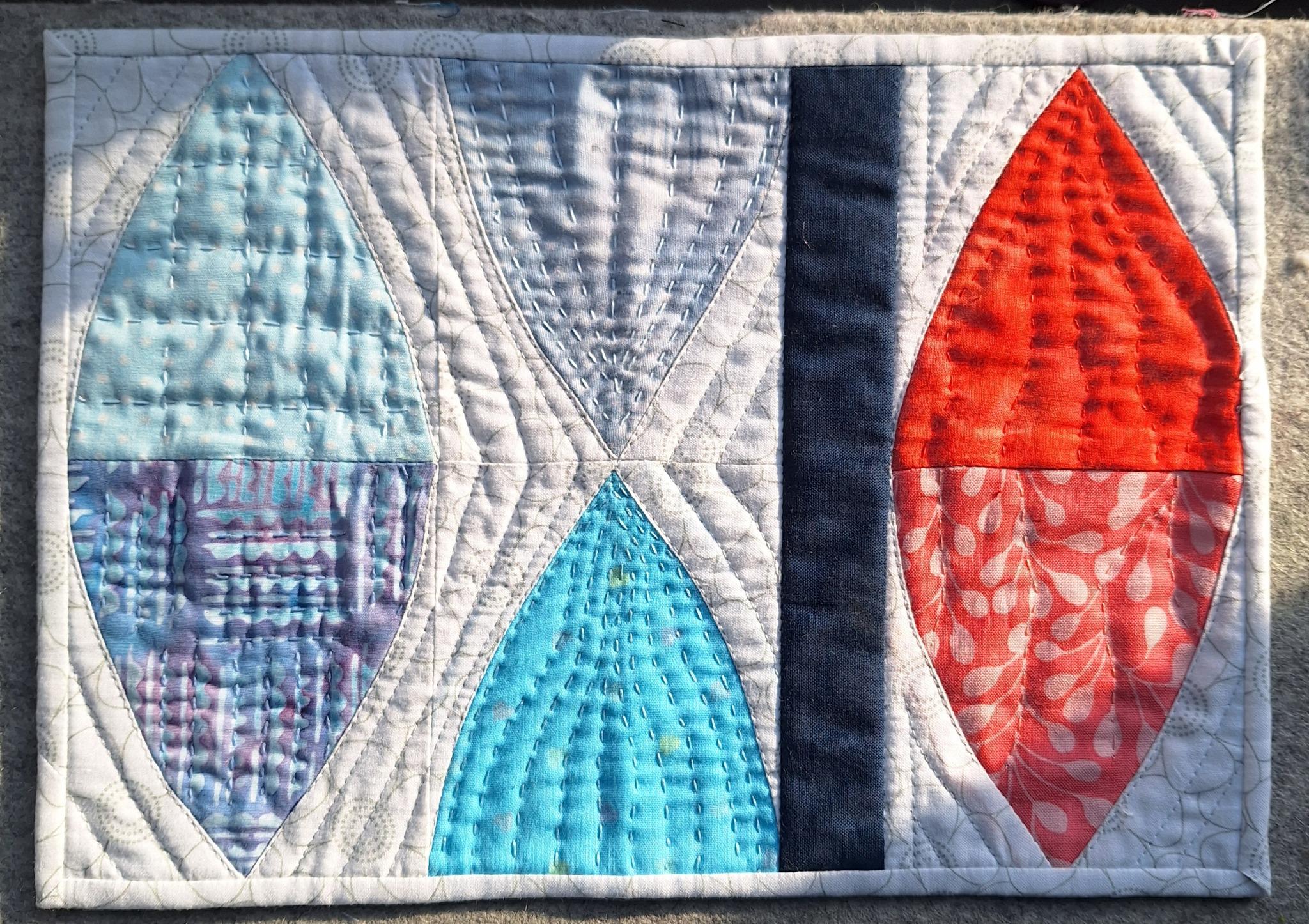 A rectangular quilted placemat with a white background and a black border. The placemat features four teardrop-shaped pieces of fabric in blue, red, and purple, arranged in a symmetrical pattern. The background is quilted with an echoing pattern.