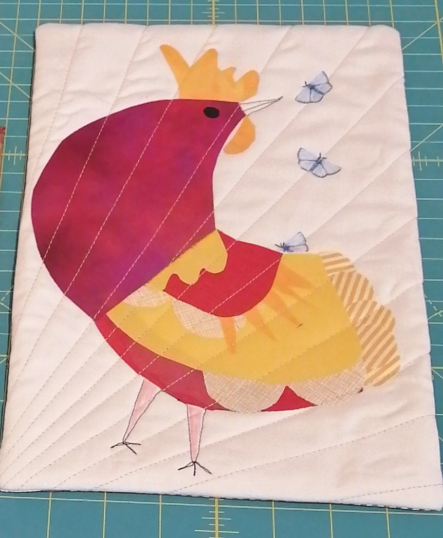 A rectangular quilted placemat with a white background. The placemat features a colourful rooster with a red body, yellow tail feathers, and a gold crown. Three small, blue butterflies are flying around the rooster. The background is quilted with radiating lines originating from the lower left corner..