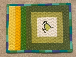 A rectangular quilted placemat with a green background. The placemat features a small, embroidered bird in the centre, surrounded by several squares of green fabric. There is a vertical stripe of yellow fabric on the left side. The placemat is framed by a border of green, blue, and yellow fabric.