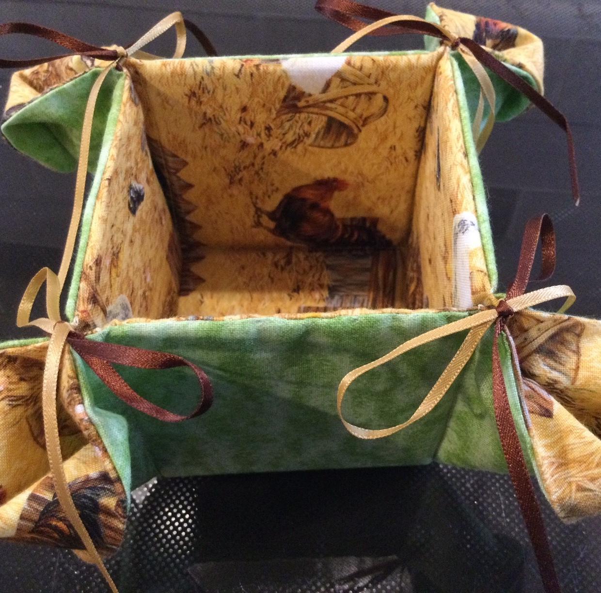 A fabric basket with a green exterior and a yellow interior. The interior features a pattern of chickens and a barn. The basket has four ribbon ties, two brown and two yellow, to keep it closed.