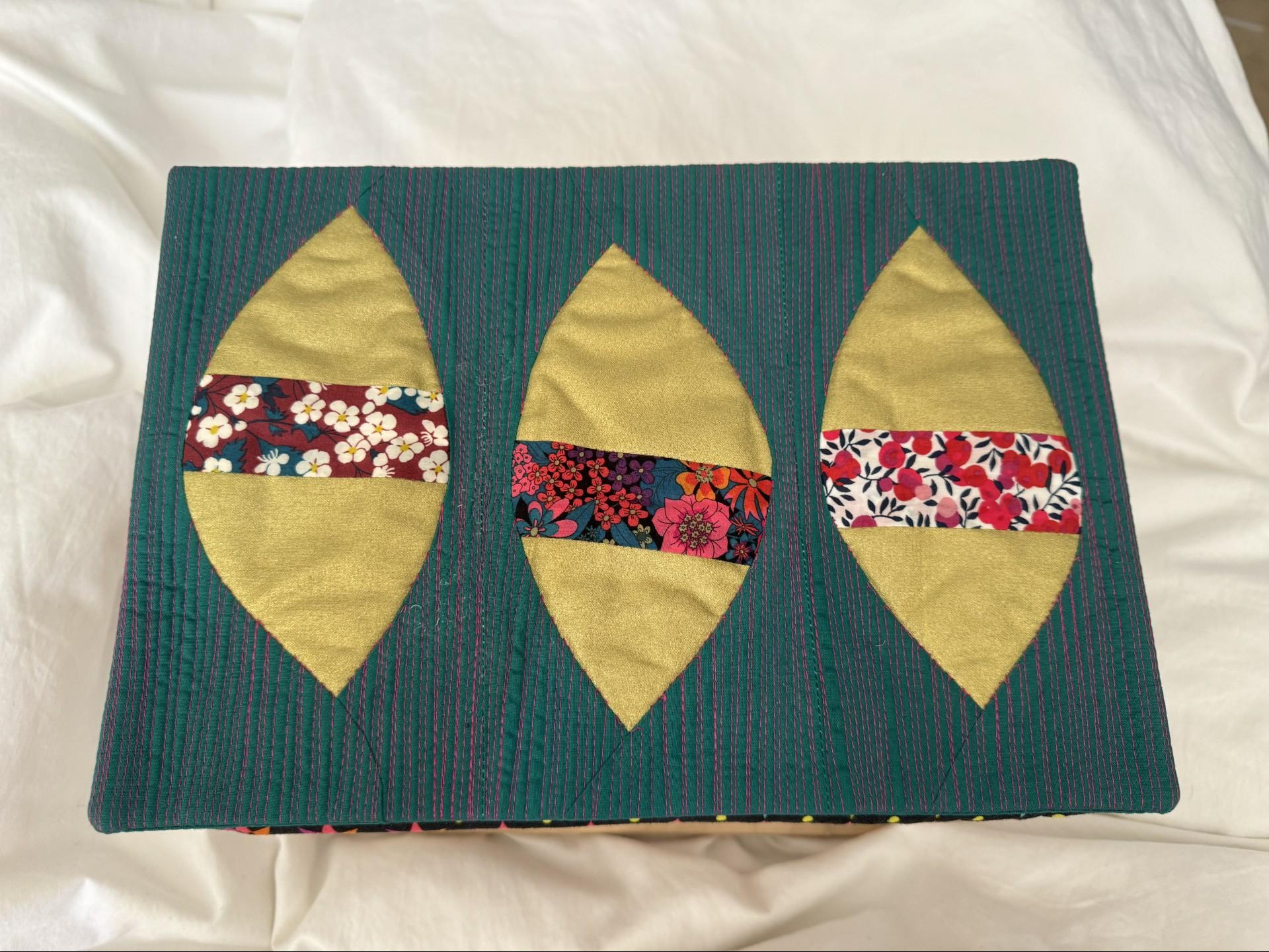 A rectangular quilted placemat with a teal background. The placemat features three overlapping, almond-shaped patches of yellow fabric. Each yellow patch has a horizontal band of colourful fabric with floral patterns. The background is quilted with red vertical lines.