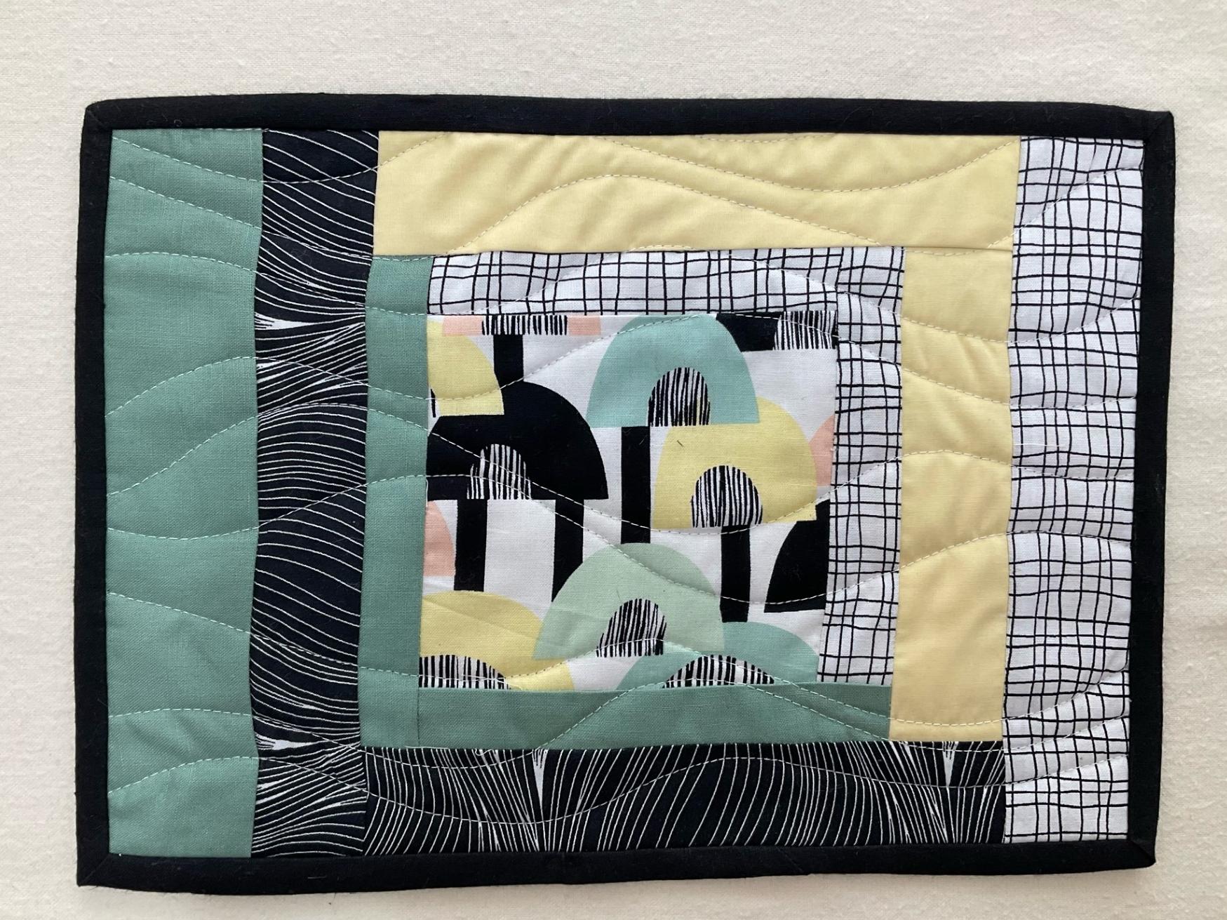 A rectangular quilted placemat with a black border. The placemat features a nested square design with a central square containing abstract shapes in black, white, green, and pink. The surrounding squares have different patterns, including waves, grids, and dots. The background is quilted with a wavy line pattern.