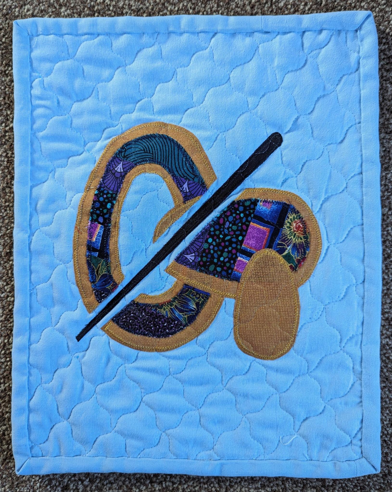 A square quilted placemat with a light blue background. The placemat features a stylised paintbrush, palette and egg, created using colourful fabric pieces. The background is quilted with a curved interlocking tiled pattern.