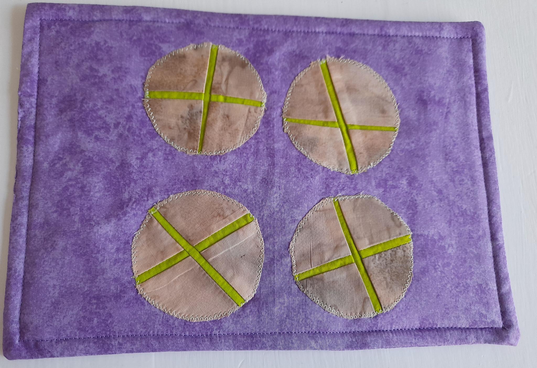 A rectangular quilted placemat with a purple background. The placemat features four circular shapes, each with a green cross stitched across it. The circles are made from a fabric with a textured, almost marbled appearance.