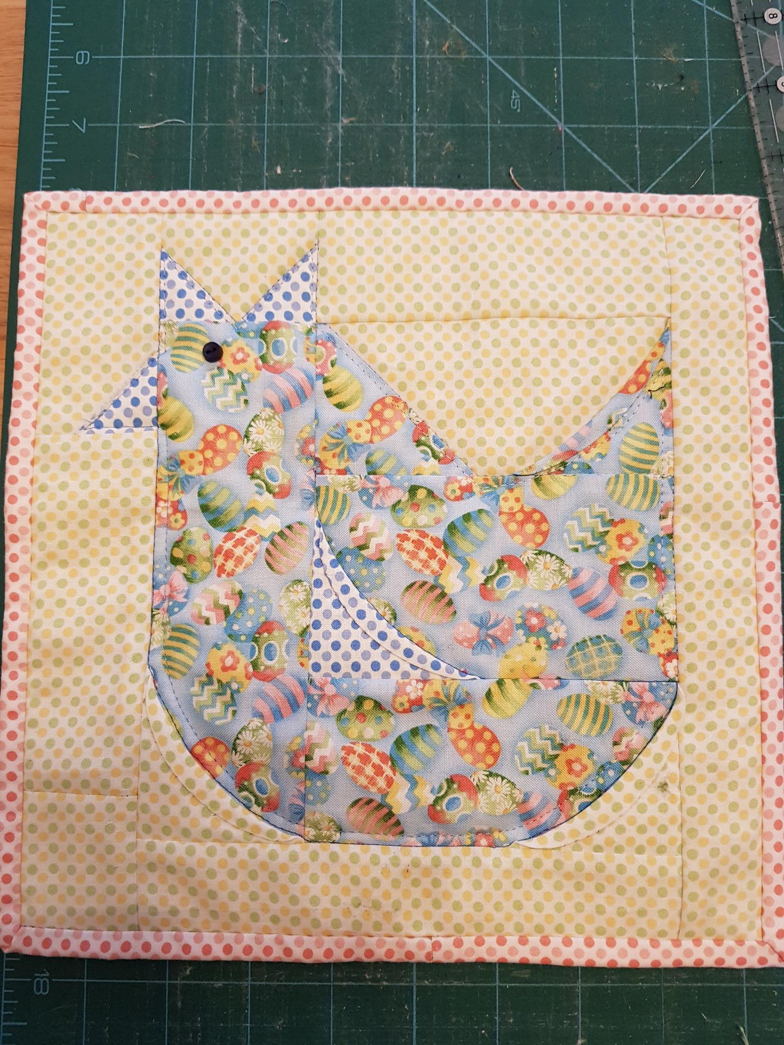 A rectangular quilted placemat with a yellow background and a pink spotted border. The placemat features a colourful chicken, made up of various fabric pieces with patterns of Easter eggs.