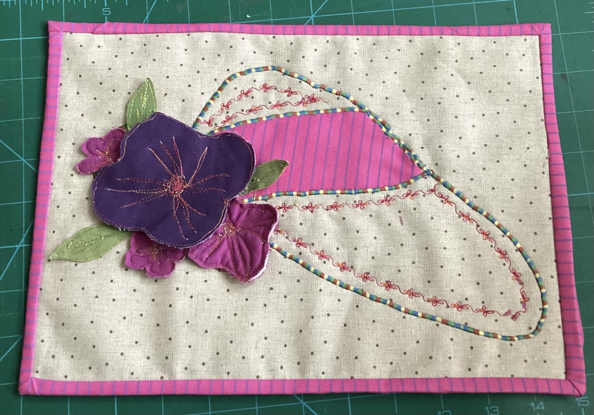 A rectangular quilted placemat with a pink border. The placemat features a large, pink flower with a purple centre, surrounded by smaller pink flowers. The flowers are appliqu&eacute;d onto a beige background with a dotted pattern.