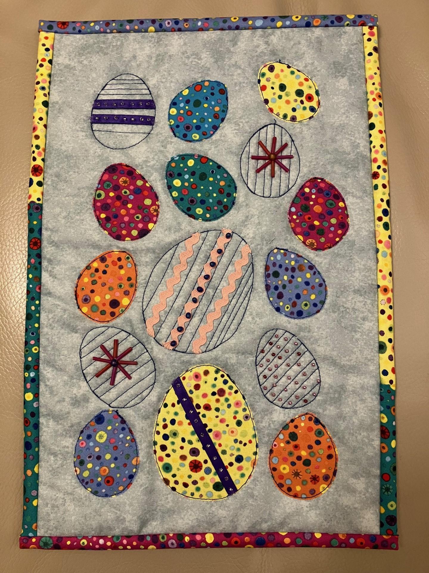A rectangular quilted placemat with a grey background and a colourful border. The placemat features multiple colourful Easter eggs of various sizes, each decorated with different patterns like stripes, dots, and stars. 