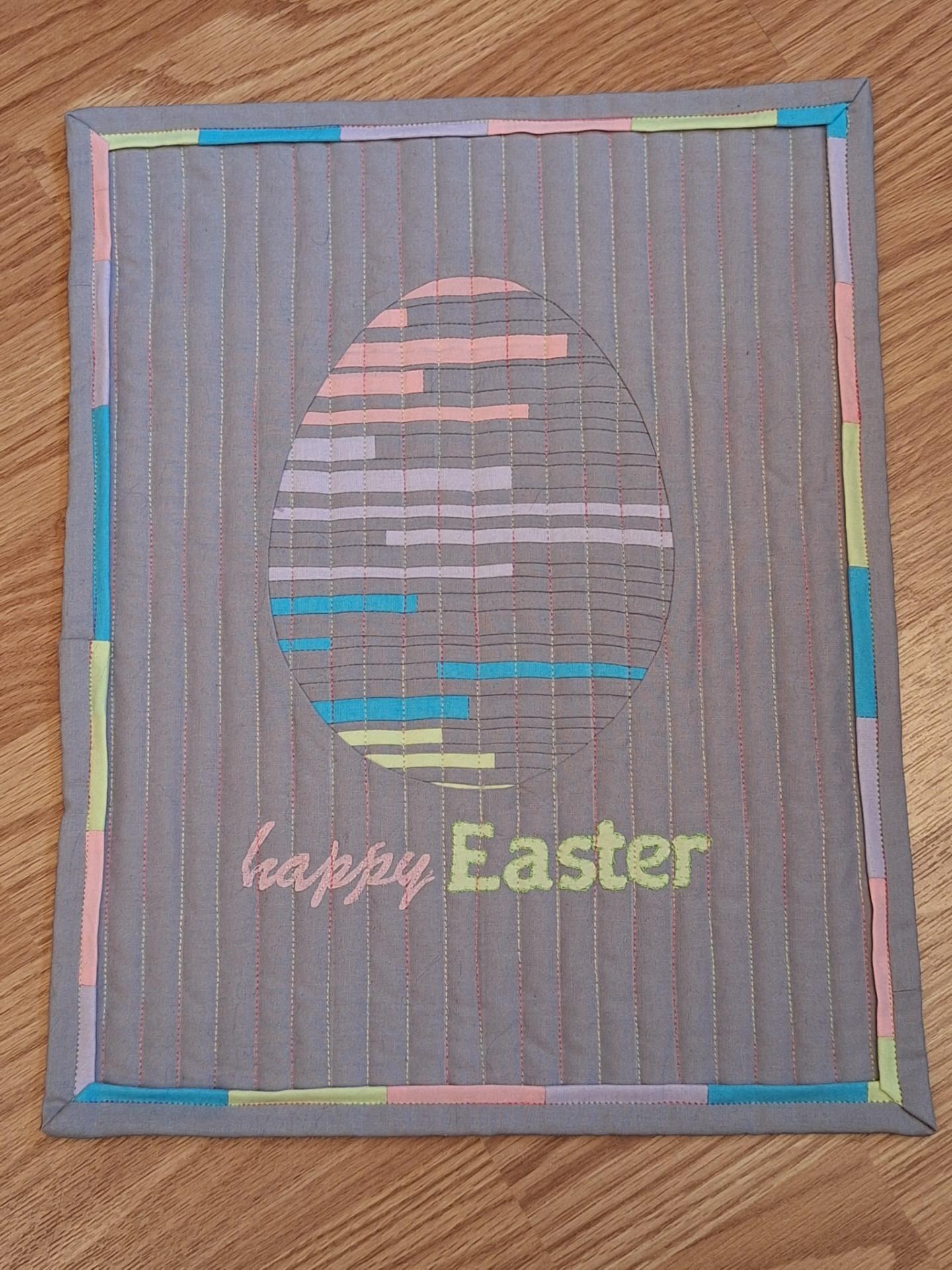 A square quilted placemat with a grey background and a colourful border. The quilt features a large, colourful Easter egg in the centre, made up of horizontal stripes in various colours. The words "Happy Easter" are embroidered below the egg. The background is quilted with vertical lines.