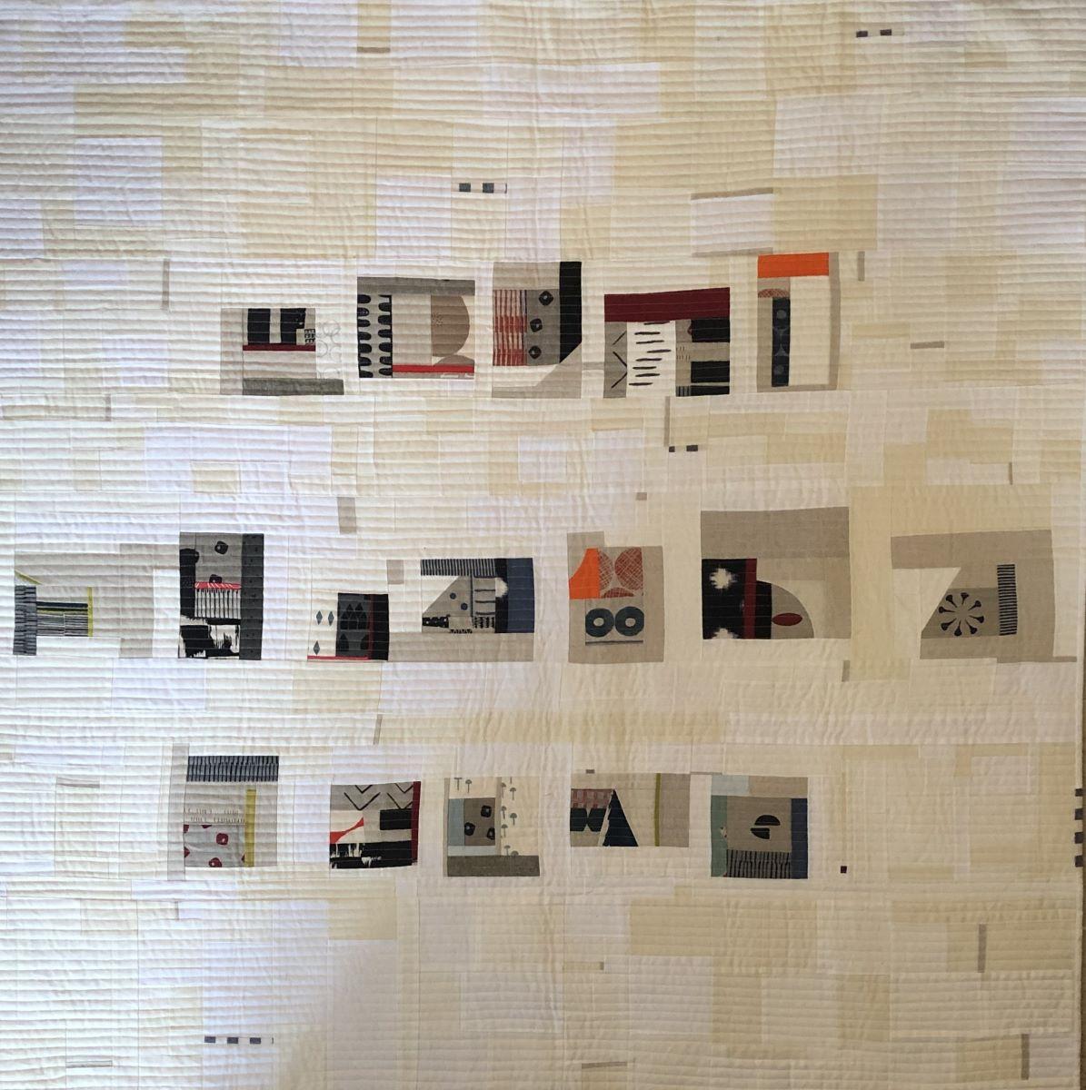 A large, abstract quilt with a predominantly white background. The quilt is composed of various shapes and patterns in black, gray, and pops of color like red and orange. There are geometric shapes, organic forms, and textural elements, creating a complex and visually interesting composition. The quilt appears to be made up of numerous smaller pieces of fabric sewn together.