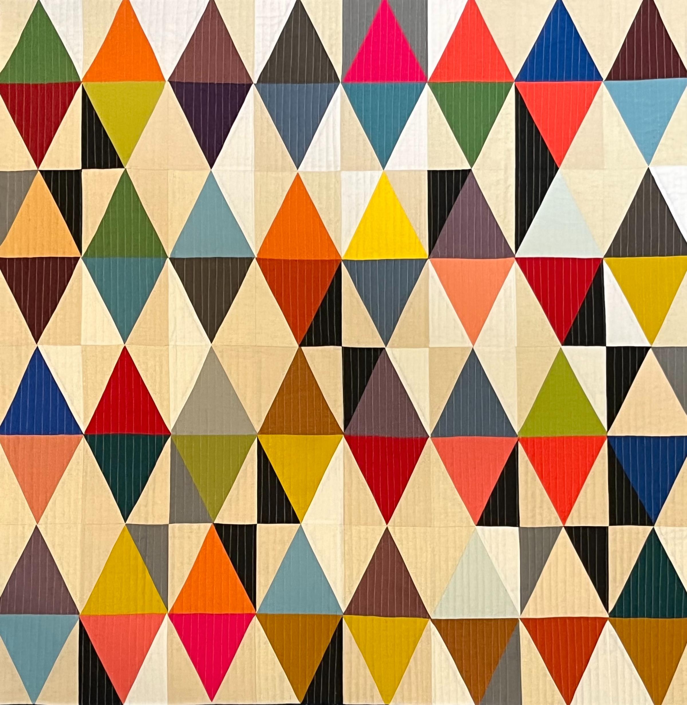 A large, abstract quilt composed of a repeating pattern of triangles in various colors, including red, pink, blue, green, yellow, and gray. The triangles are arranged in a grid-like pattern, creating a dynamic and visually interesting composition.