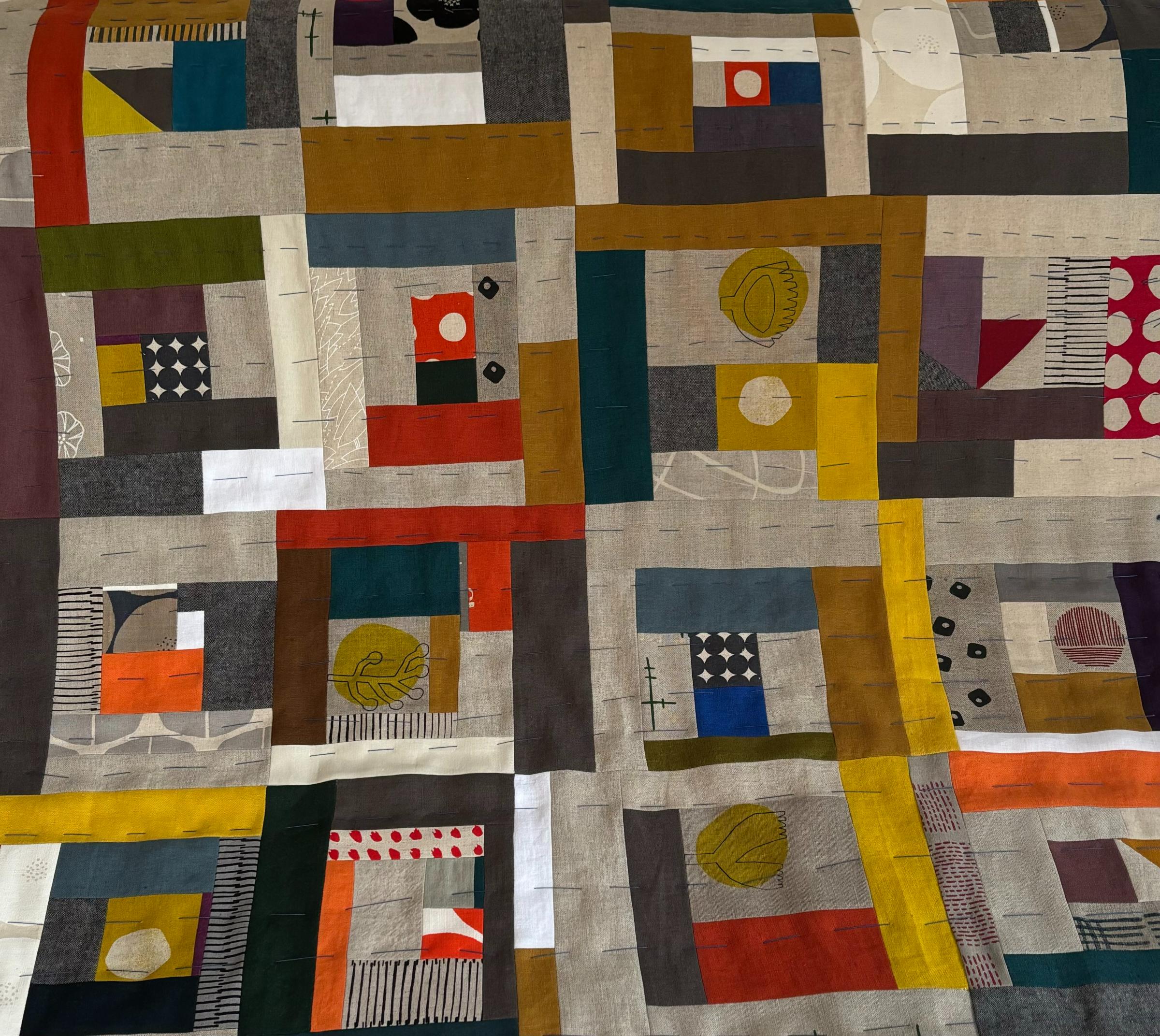 A large, abstract quilt with a predominantly neutral color palette of gray, brown, and white. The quilt is composed of various shapes and patterns, including squares, rectangles, and circles, in different sizes and colors. There are also pops of color like red, yellow, and green. The quilt appears to be made up of numerous smaller pieces of fabric sewn together.