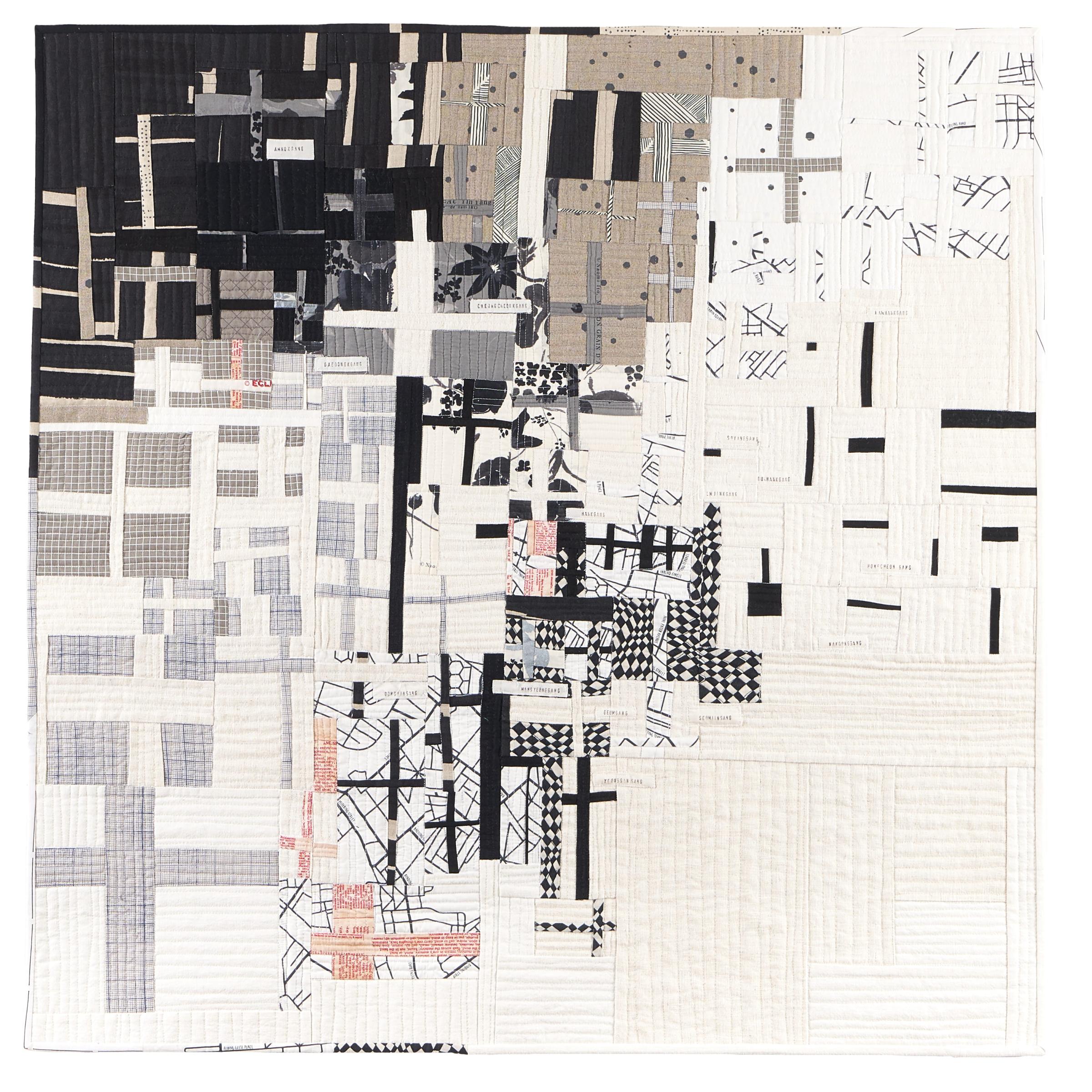 A large, abstract quilt with a predominantly white background. The quilt is composed of various shapes and patterns in black, gray, and white. There are geometric shapes, organic forms, and textural elements, creating a complex and visually interesting composition. The quilt appears to be made up of numerous smaller pieces of fabric sewn together.