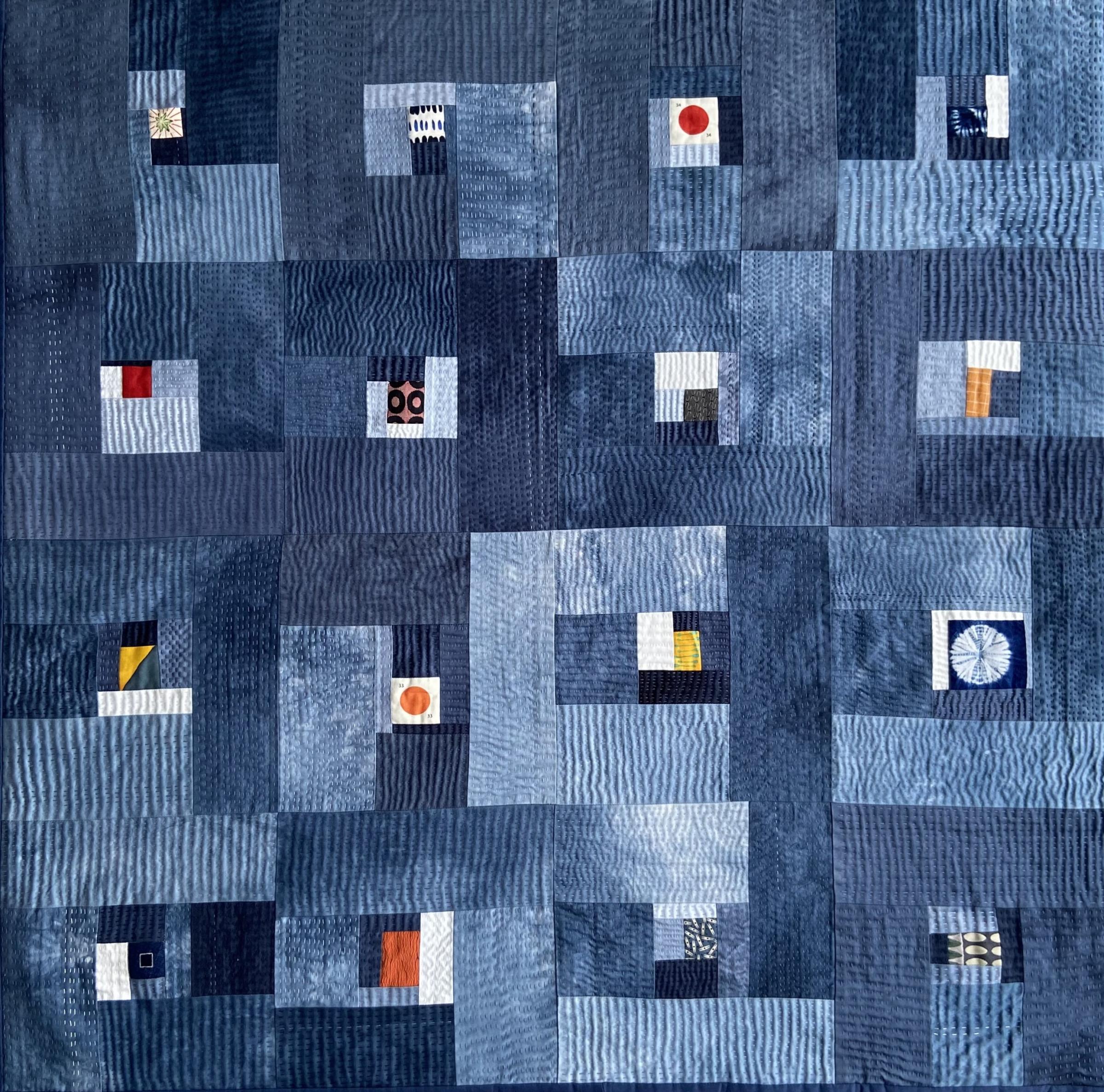 A large, abstract quilt with a predominantly blue background. The quilt is composed of various shades of blue fabric, with some squares featuring small pops of color like red, yellow, and white. The quilt appears to be made up of numerous smaller squares of fabric sewn together.